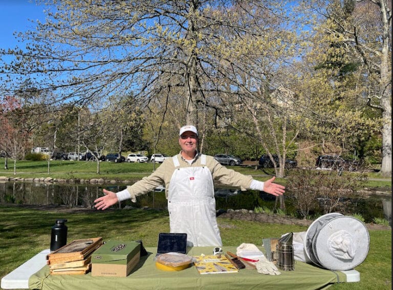 Joel Dawson in front of a park showcasing his honey materials with open arms and a great smile.