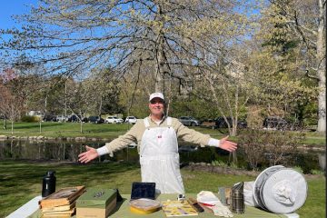 Joel Dawson in front of a park showcasing his honey materials with open arms and a great smile.